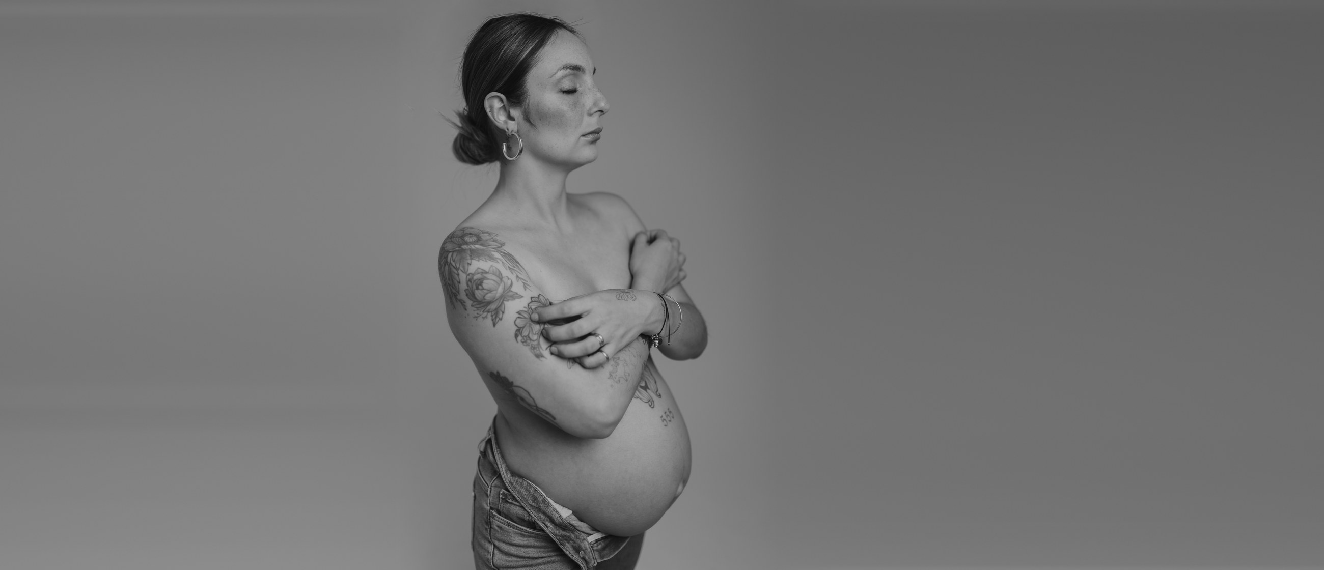 Grávida de perfil em estúdio, tatuagens no ombro e na barriga, a preto e branco