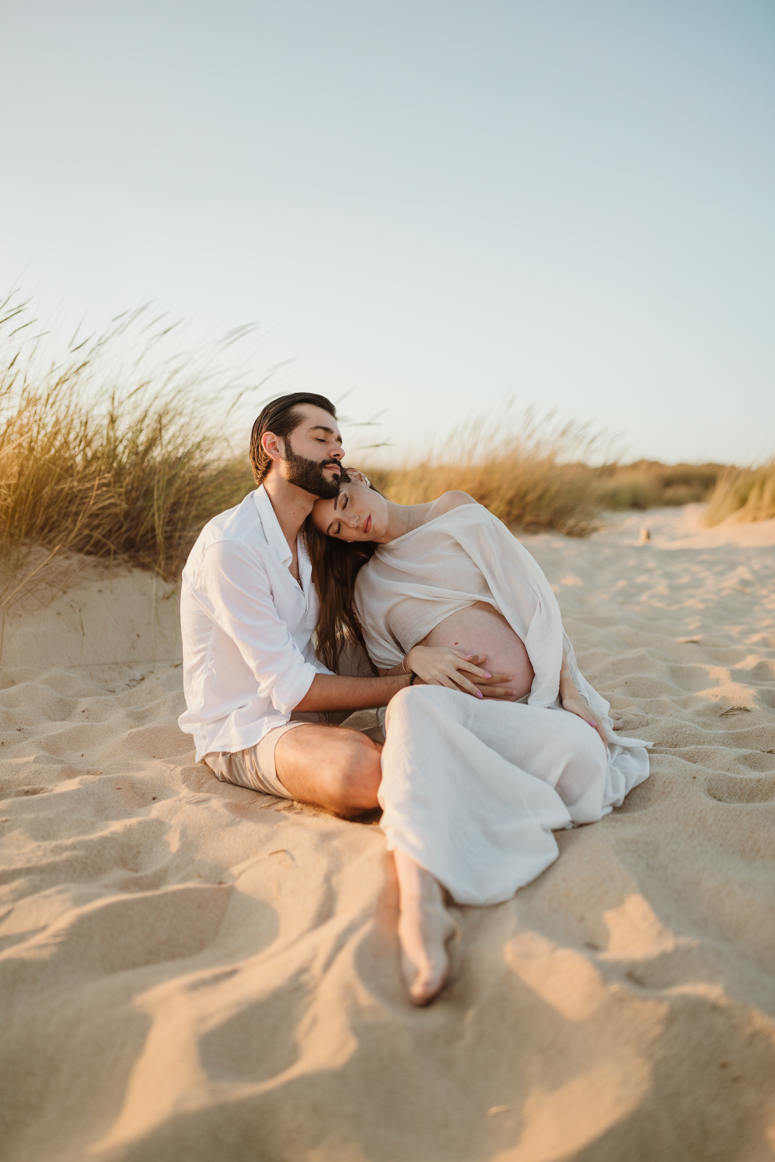 Casal grávido abraçado nas dunas, mãos na barriga, pôr do sol