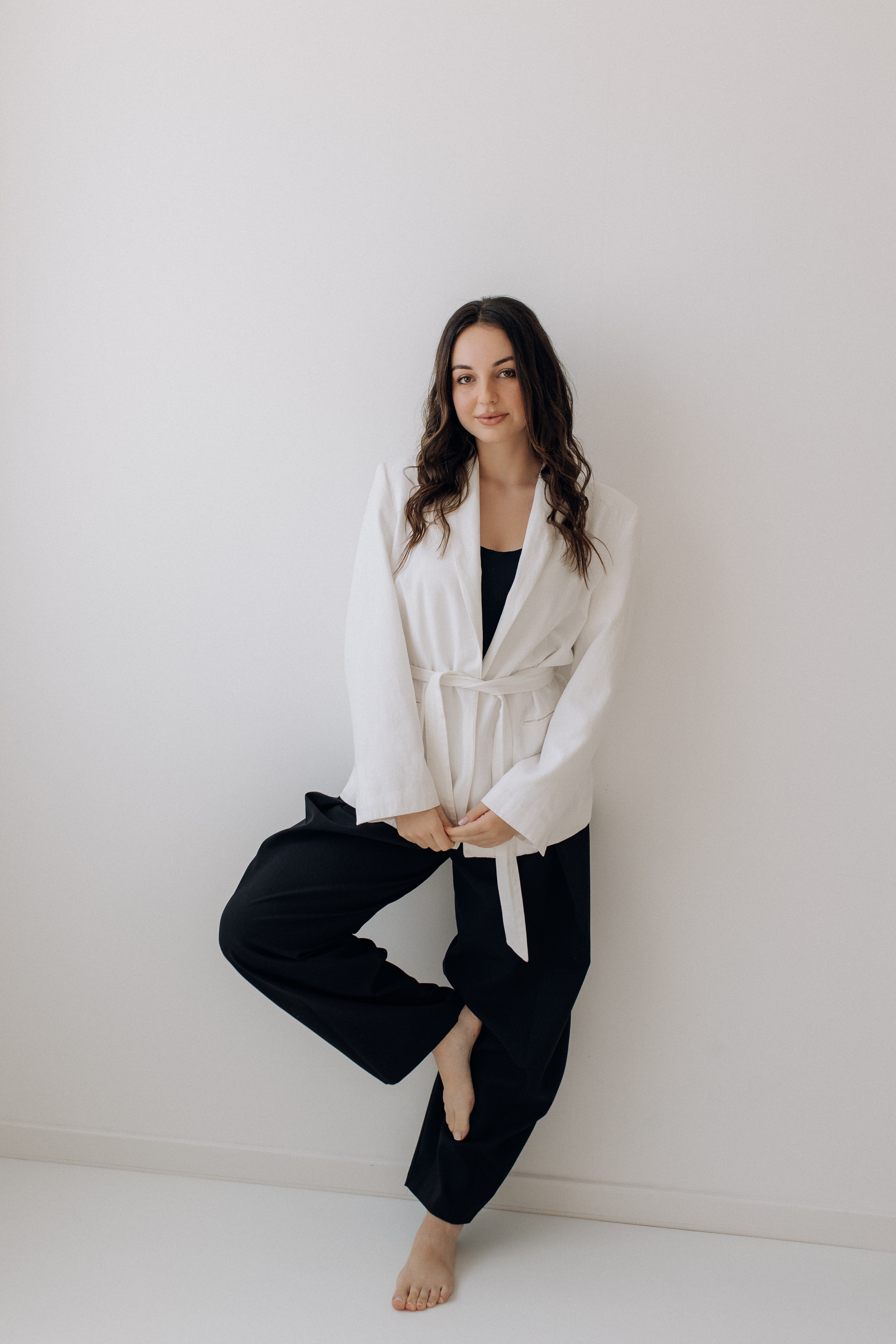 Mulher de blazer branco encostada a uma parede clara de estúdio, em luz natural