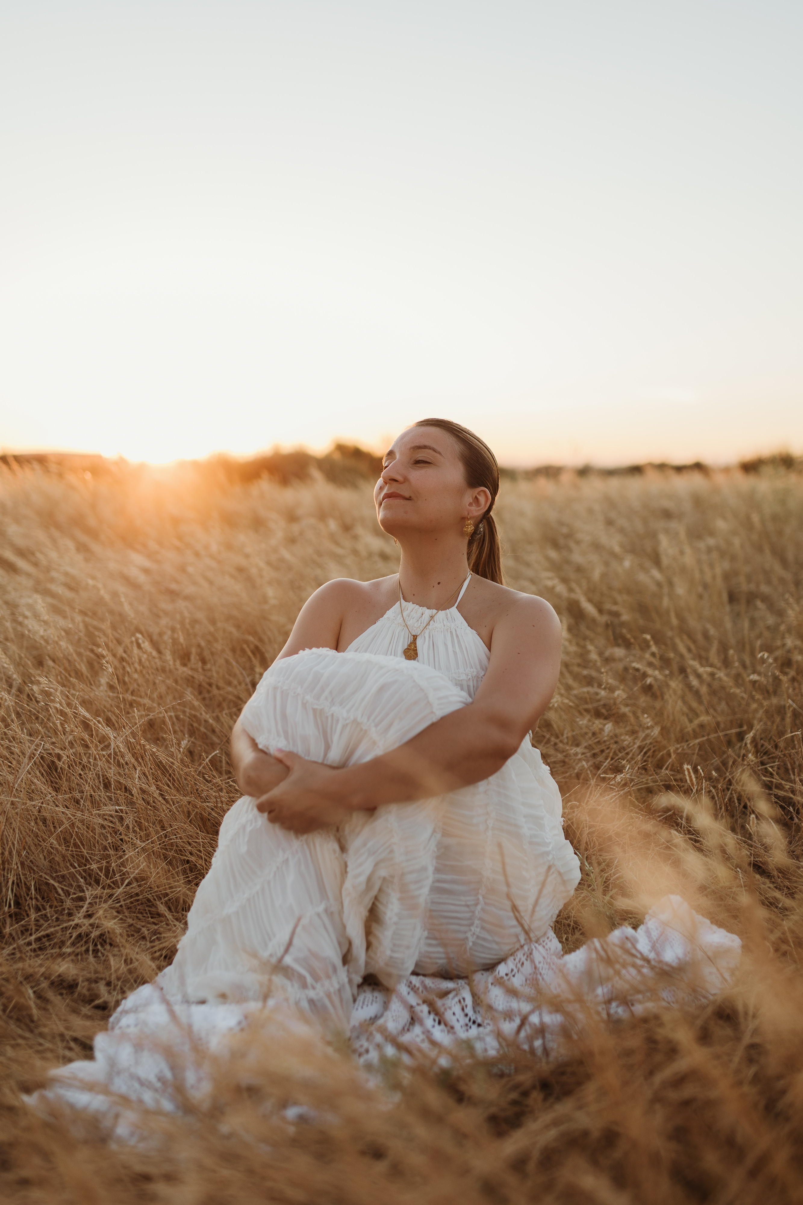 Mulher sentada no campo, de olhos fechados, contraluz do pôr do sol