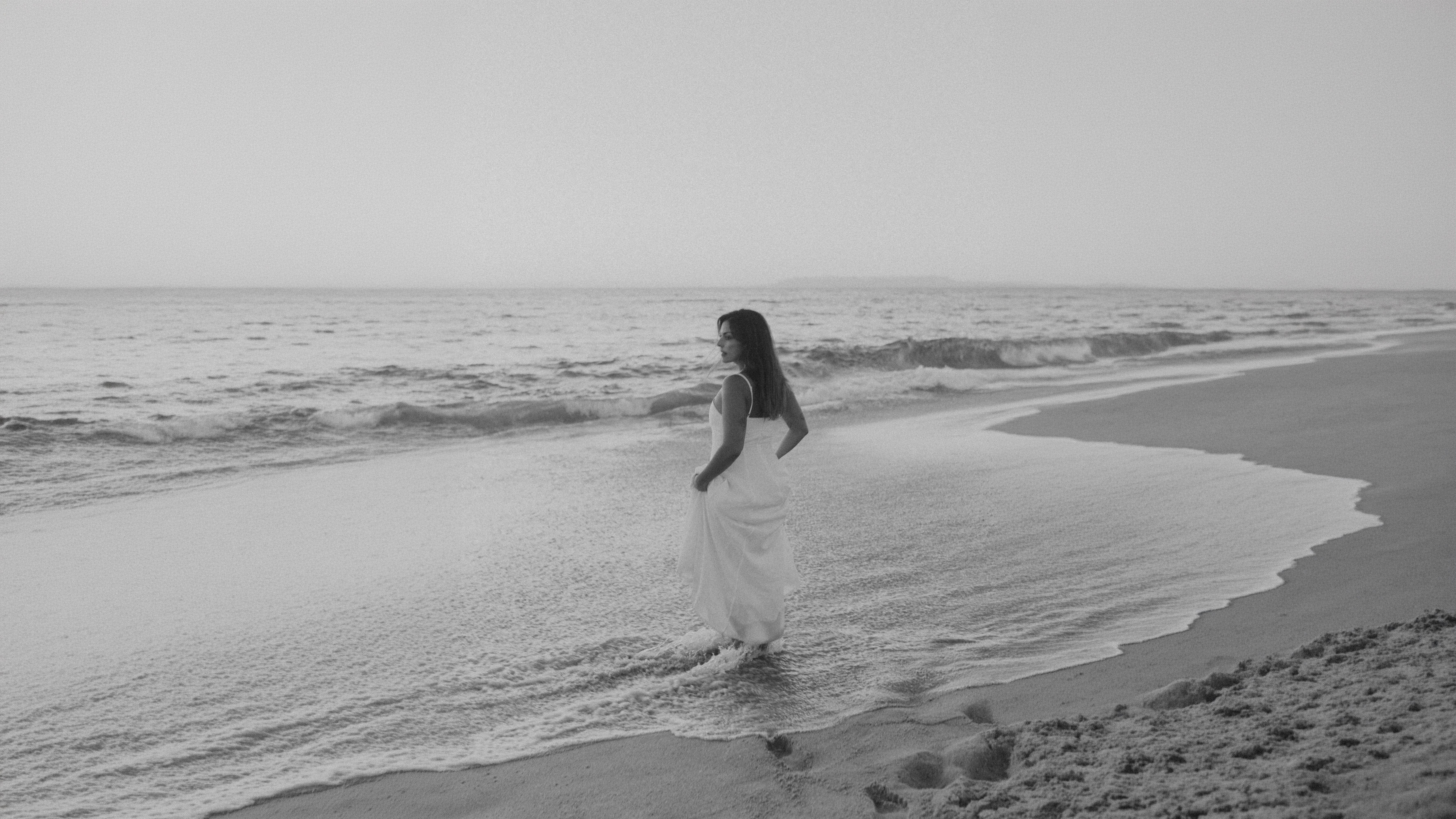 Mulher de vestido branco na rebentação, a olhar para o mar
