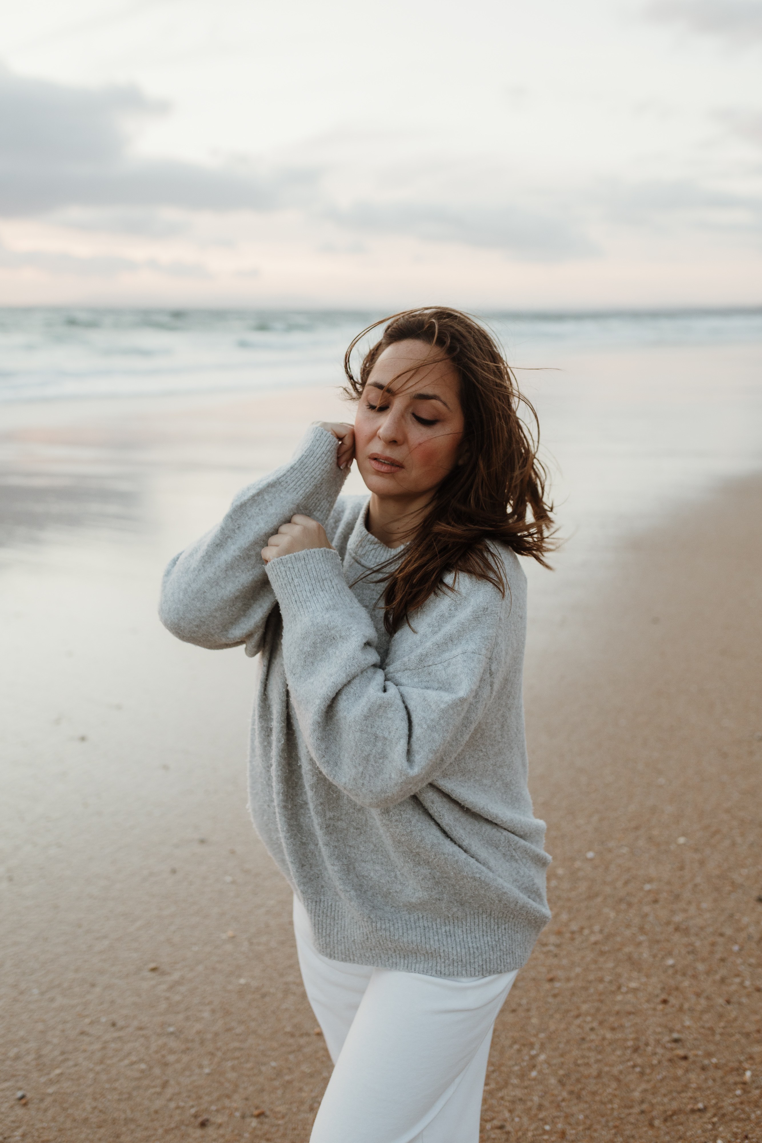 Mulher de camisola cinza na praia, cabelo ao vento, olhos fechados