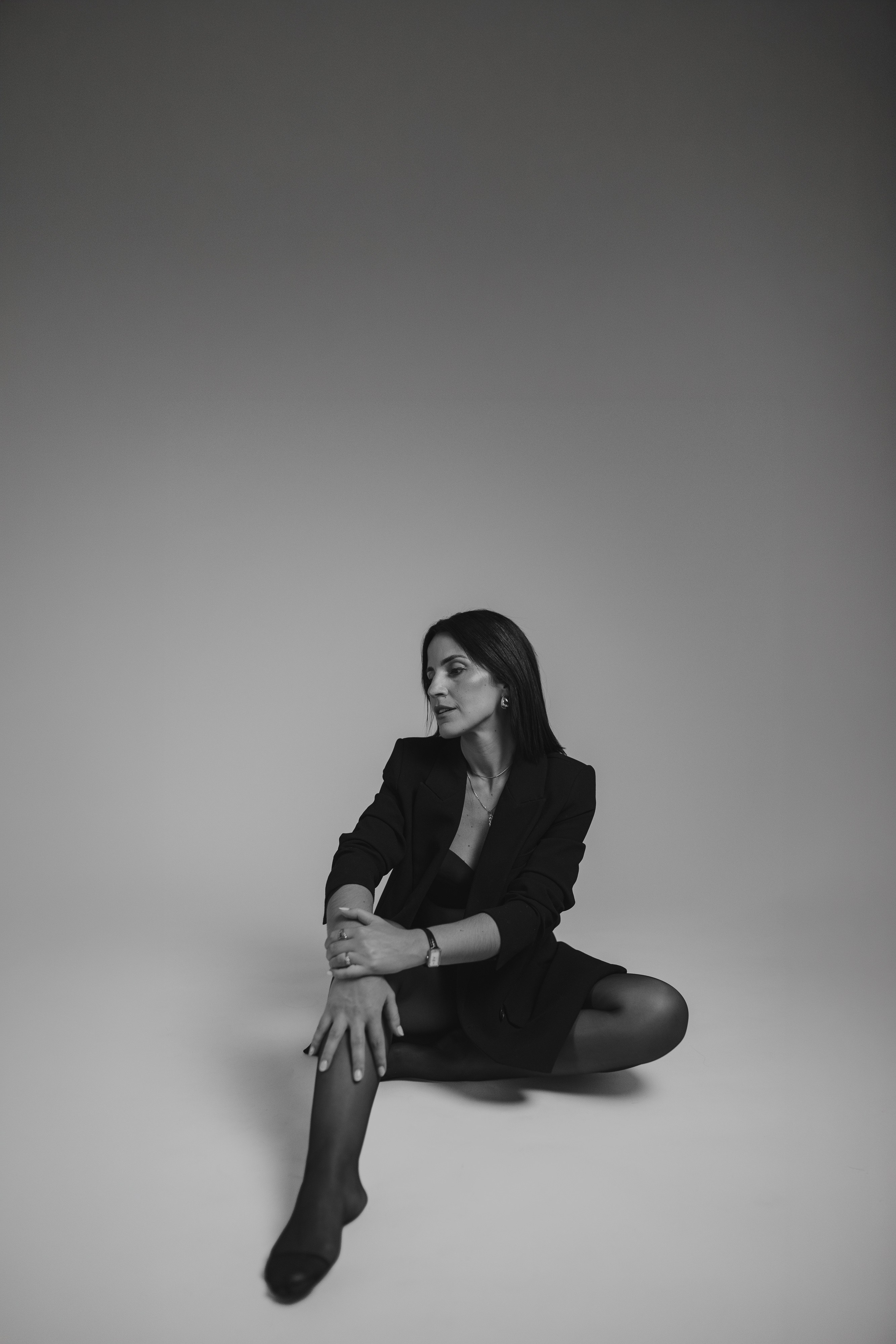 Mulher de blazer preto sentada no chão do estúdio, fundo cinzento