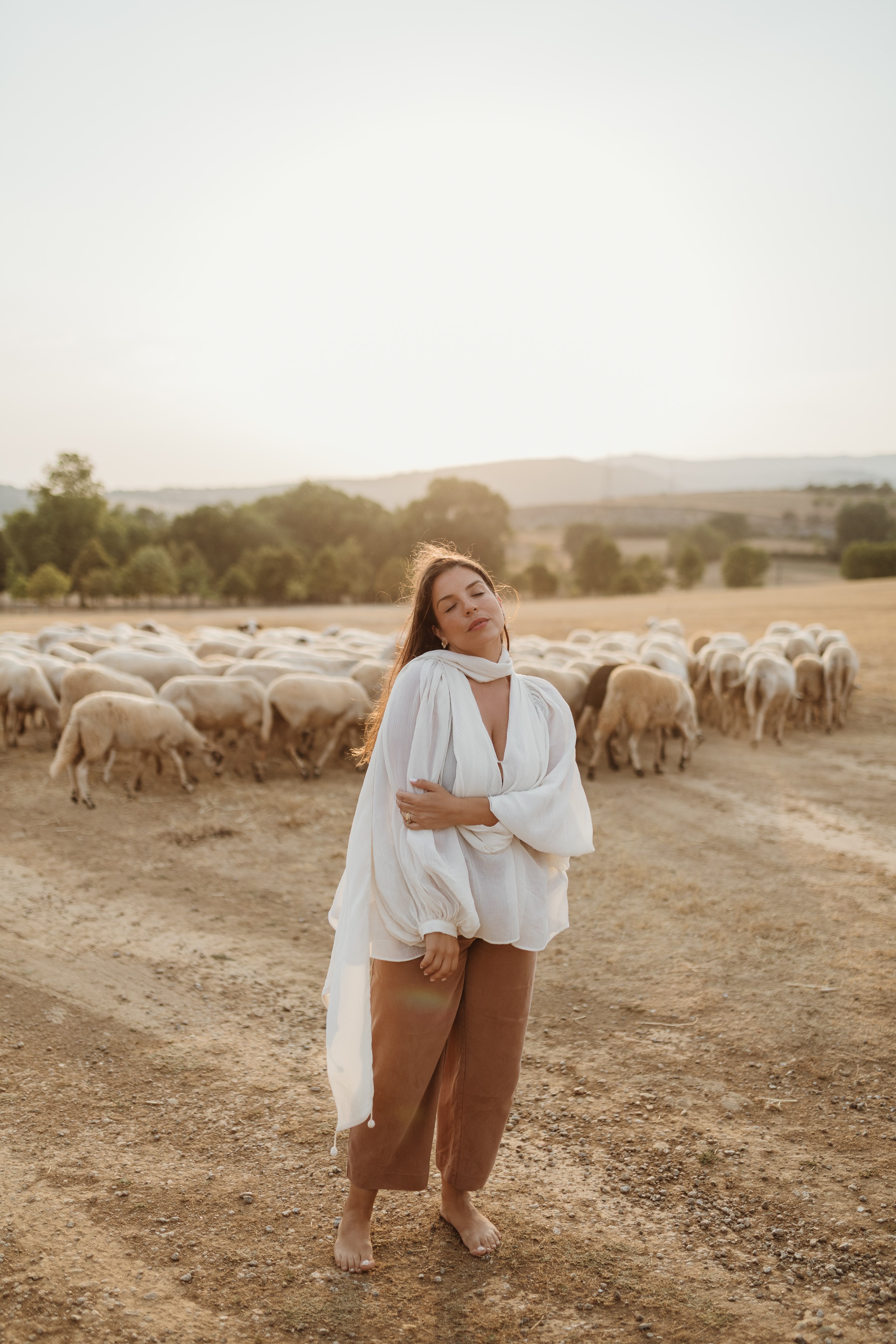 Mulher de olhos fechados junto a um rebanho de ovelhas, ao entardecer
